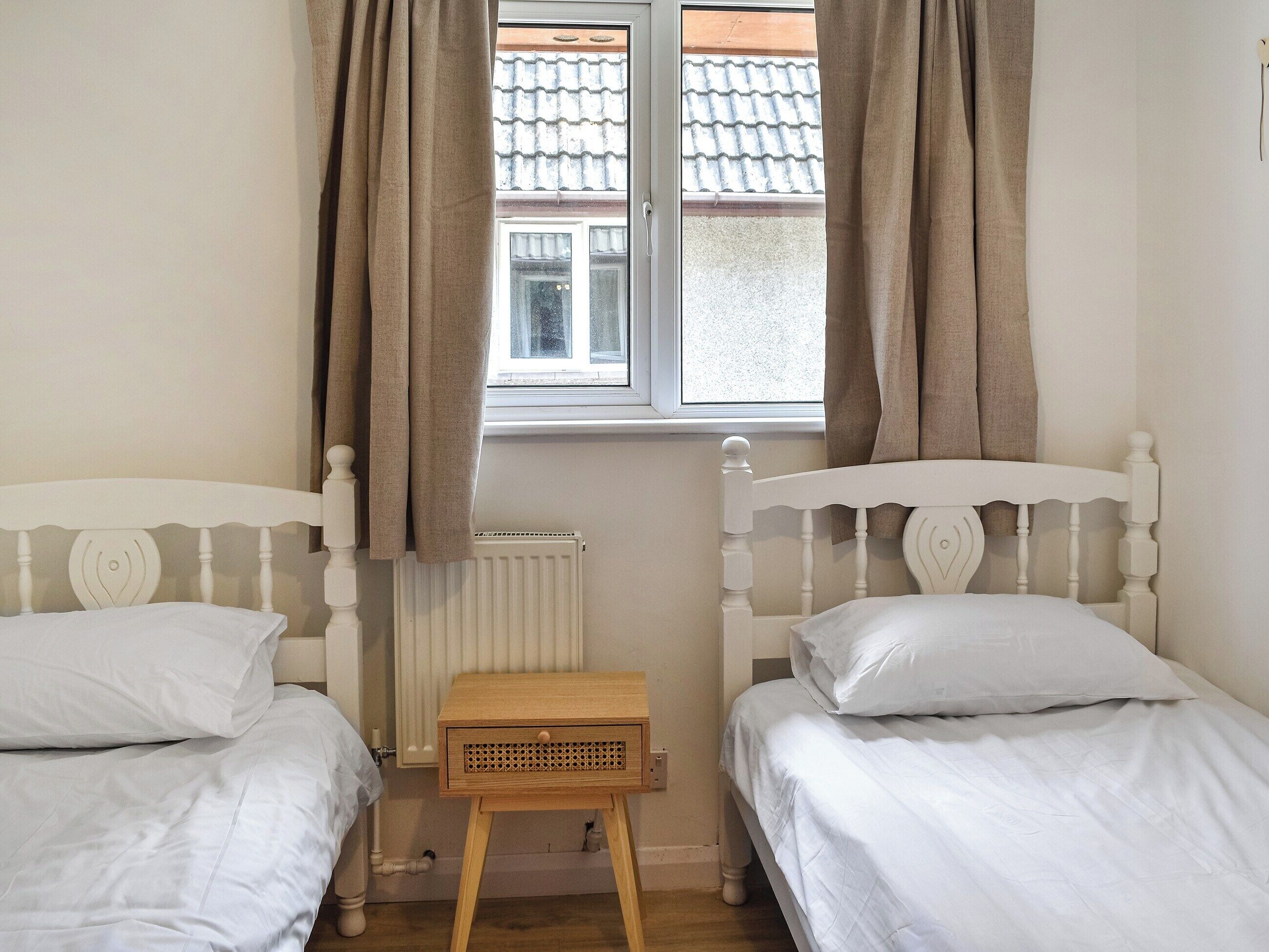 A room with two white beds, a wooden nightstand, a window with curtains, and a radiator.
