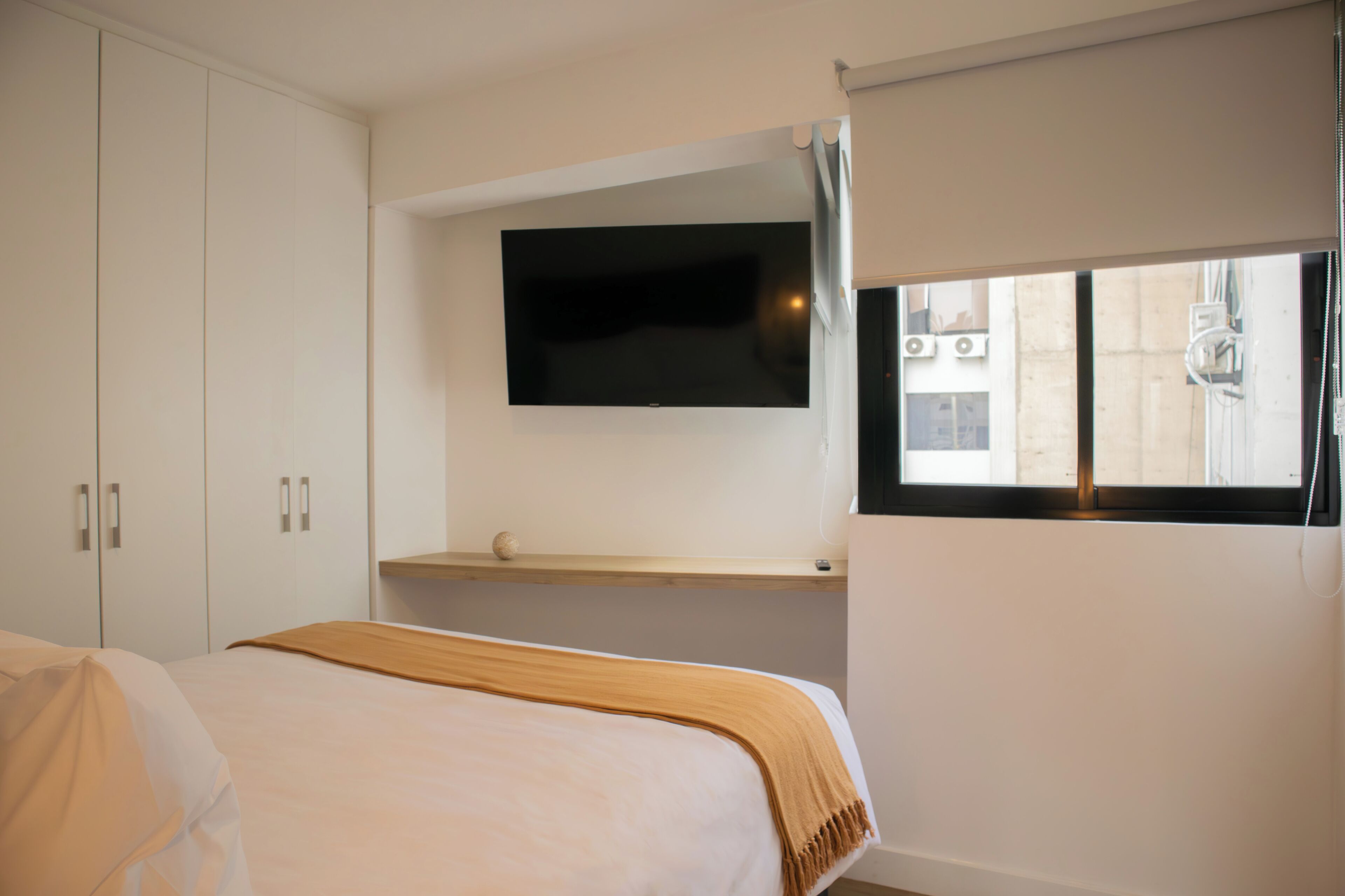 A bedroom with a bed, a TV mounted on the wall, and a window with blinds.