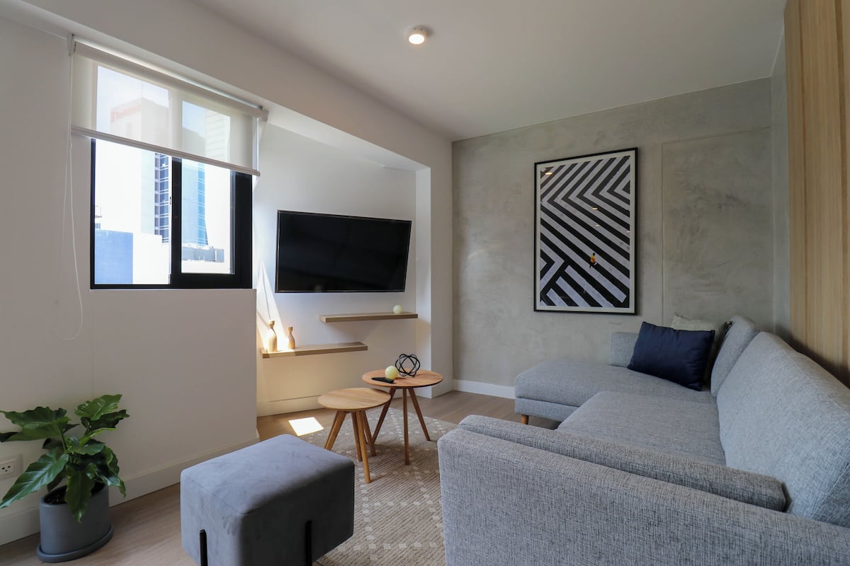 A modern living room with a grey sofa, a flat-screen TV mounted on the wall, a small wooden table, and a potted plant.