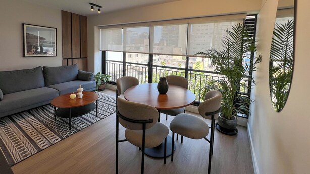 Una sala de estar moderna con sofá, mesa de centro, mesa de comedor, sillas, ventanas amplias y plantas en macetas.