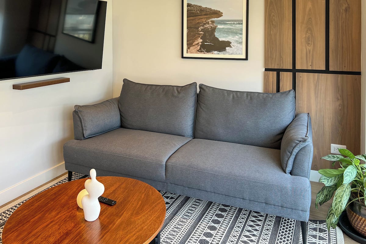 A modern living room with a grey sofa, a mounted TV, a framed coastal landscape picture, a wooden sideboard, and a patterned rug.