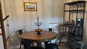 A dining area with a round wooden table, black chairs, a hanging light fixture, a framed picture on the wall, and a metal shelf with kitchen items.