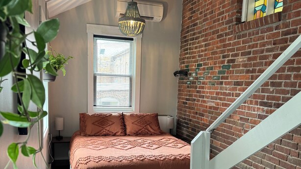 A bedroom with a bed, a hanging plant, a window with blinds, a brick wall, and a staircase.