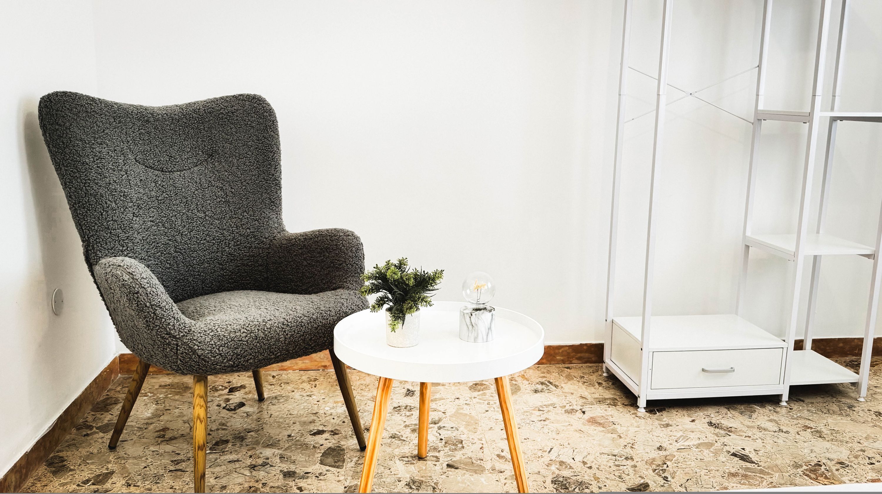 A room with a textured grey armchair, a small white table with a plant and a glass sphere, and a white shelving unit.
