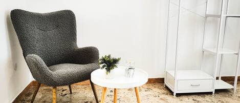 A room with a textured grey armchair, a small white table with a plant and a glass sphere, and a white shelving unit.