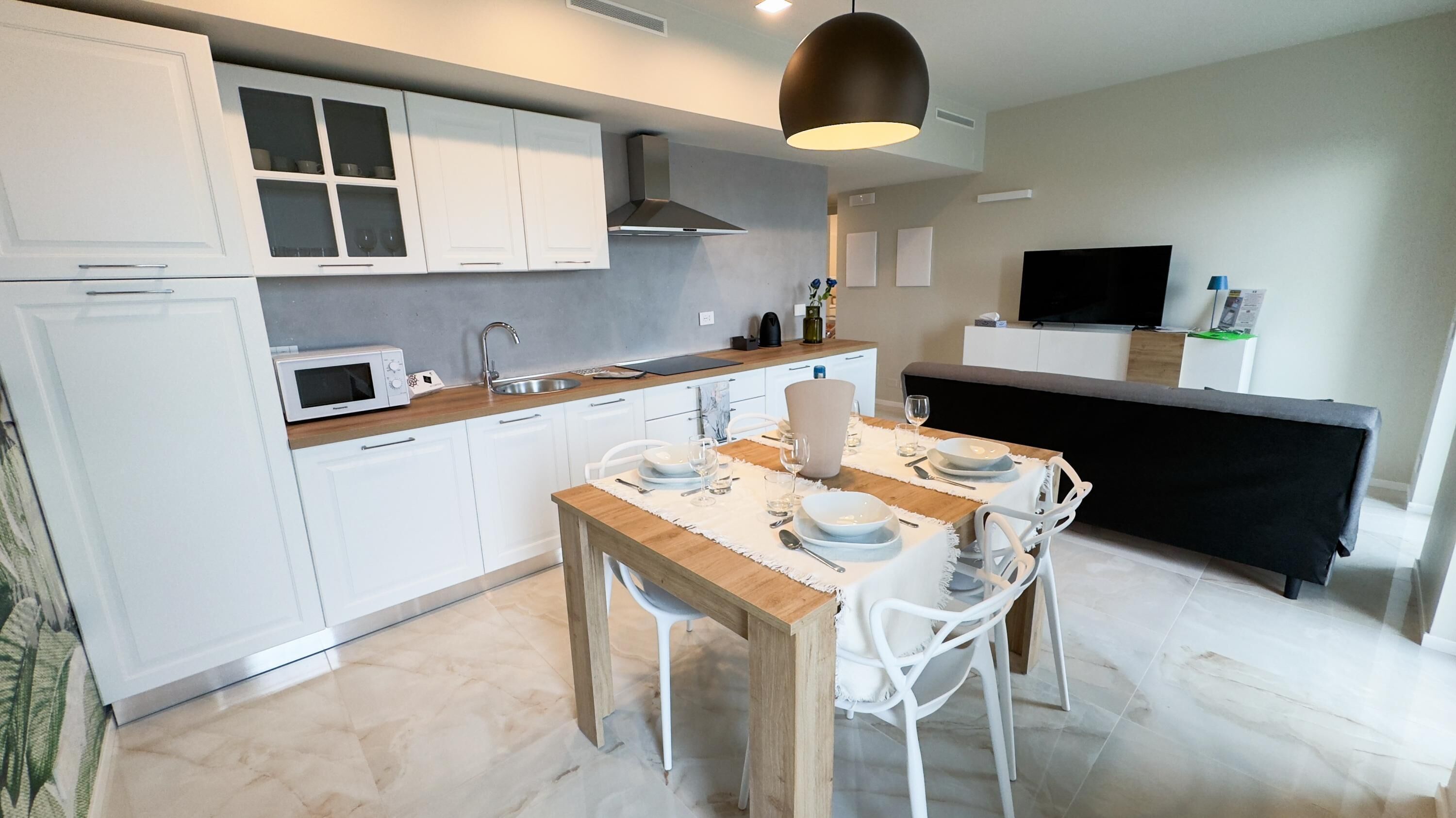 A modern kitchen with white cabinets, a wooden dining table, and a black TV on the wall.