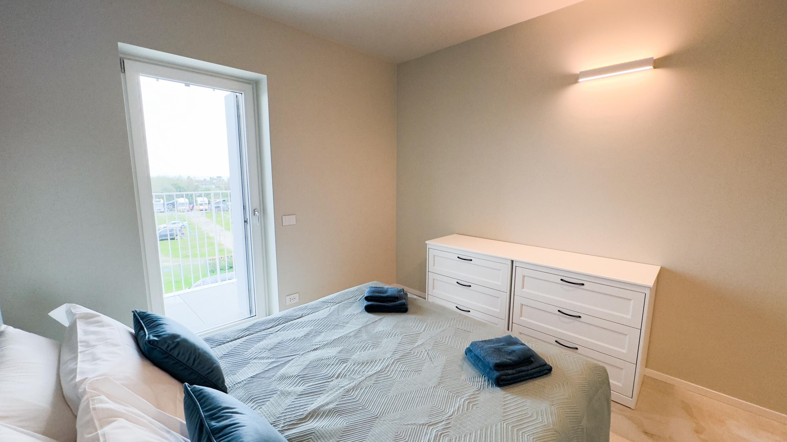Een slaapkamer met een bed, een witte dressoir en een raam met uitzicht op een parkeerplaats.