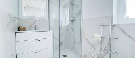 Un baño moderno con ducha de vidrio, tocador blanco con cajones y pared cubierta de mármol.