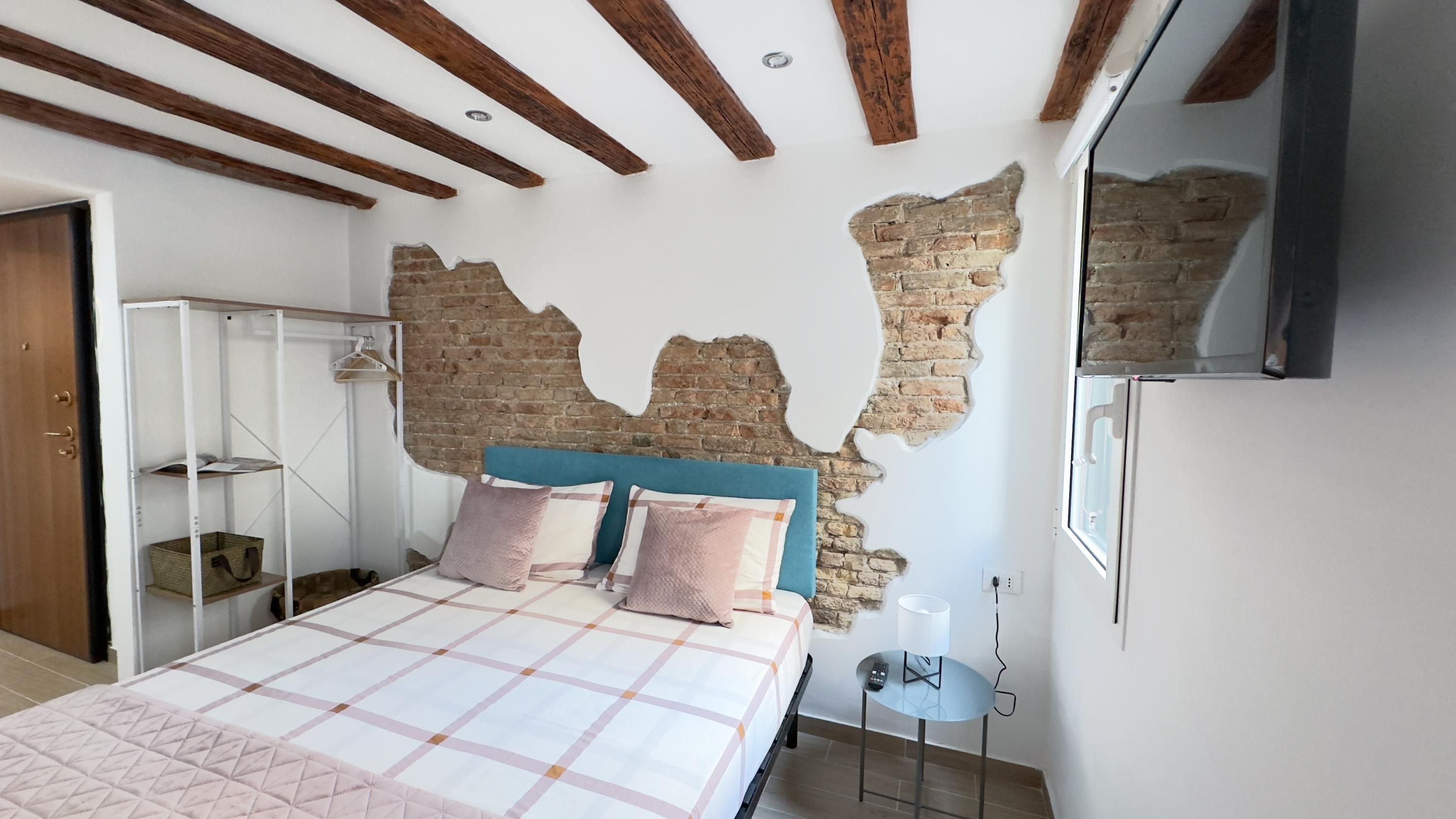 Quarto com paredes de tijolo aparente, cama com cabeceira azul, mesa de cabeceira com luminária e guarda-roupa com cabideiro.