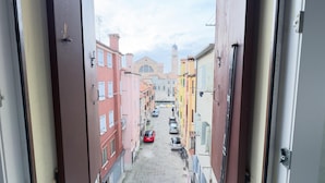Una stretta via fiancheggiata da edifici, con un'auto rossa parcheggiata al centro e una chiesa con un alto campanile in lontananza.