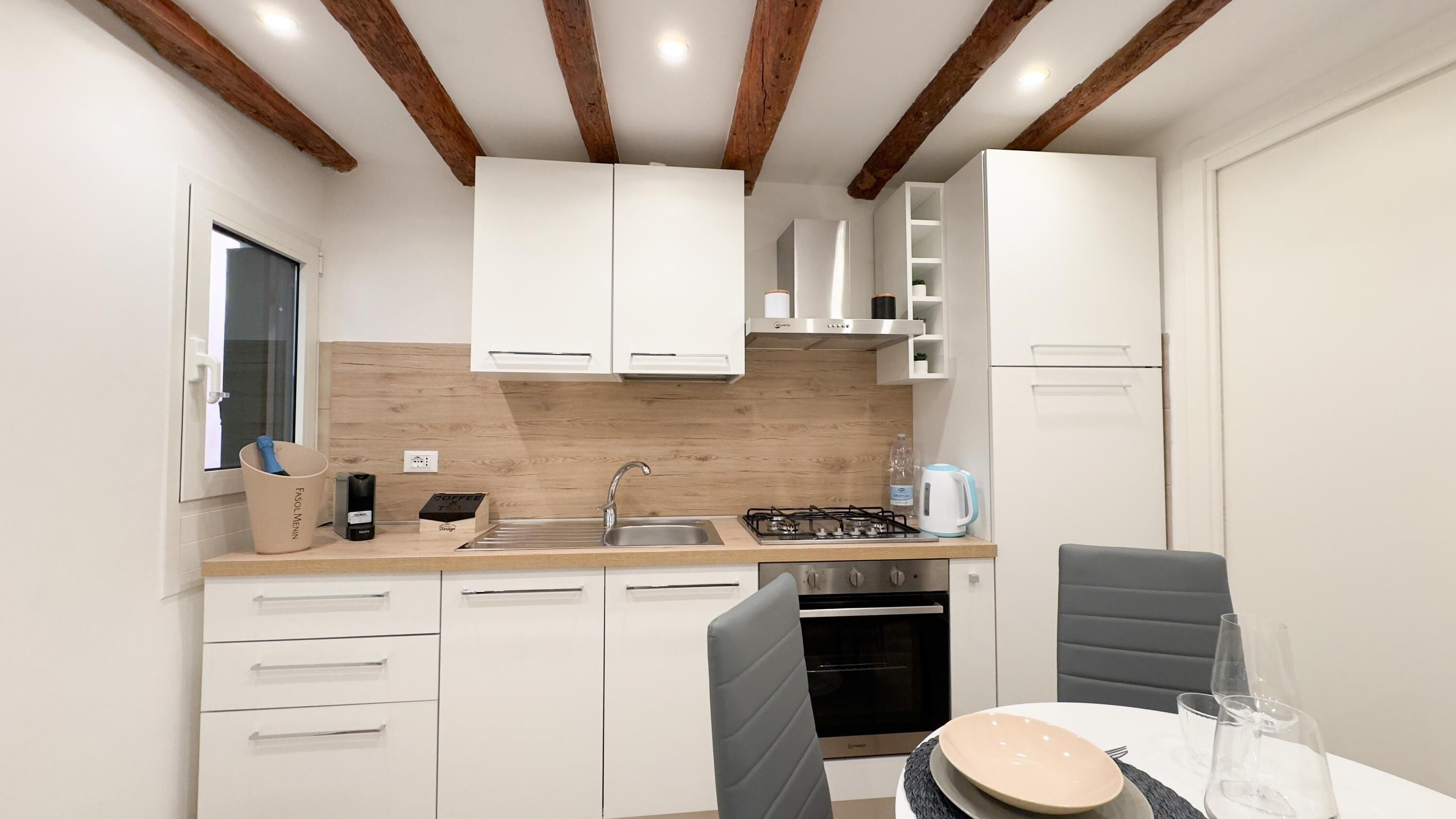 Une cuisine moderne avec des armoires blanches, un plan de travail en bois, une cuisinière à gaz et un four encastré. On y trouve un évier avec un robinet, ainsi qu’une machine à café posée sur le plan de travail.