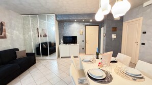 Una sala de estar moderna con un sofá negro, una mesa de comedor para dos personas y un televisor sobre un mueble blanco.