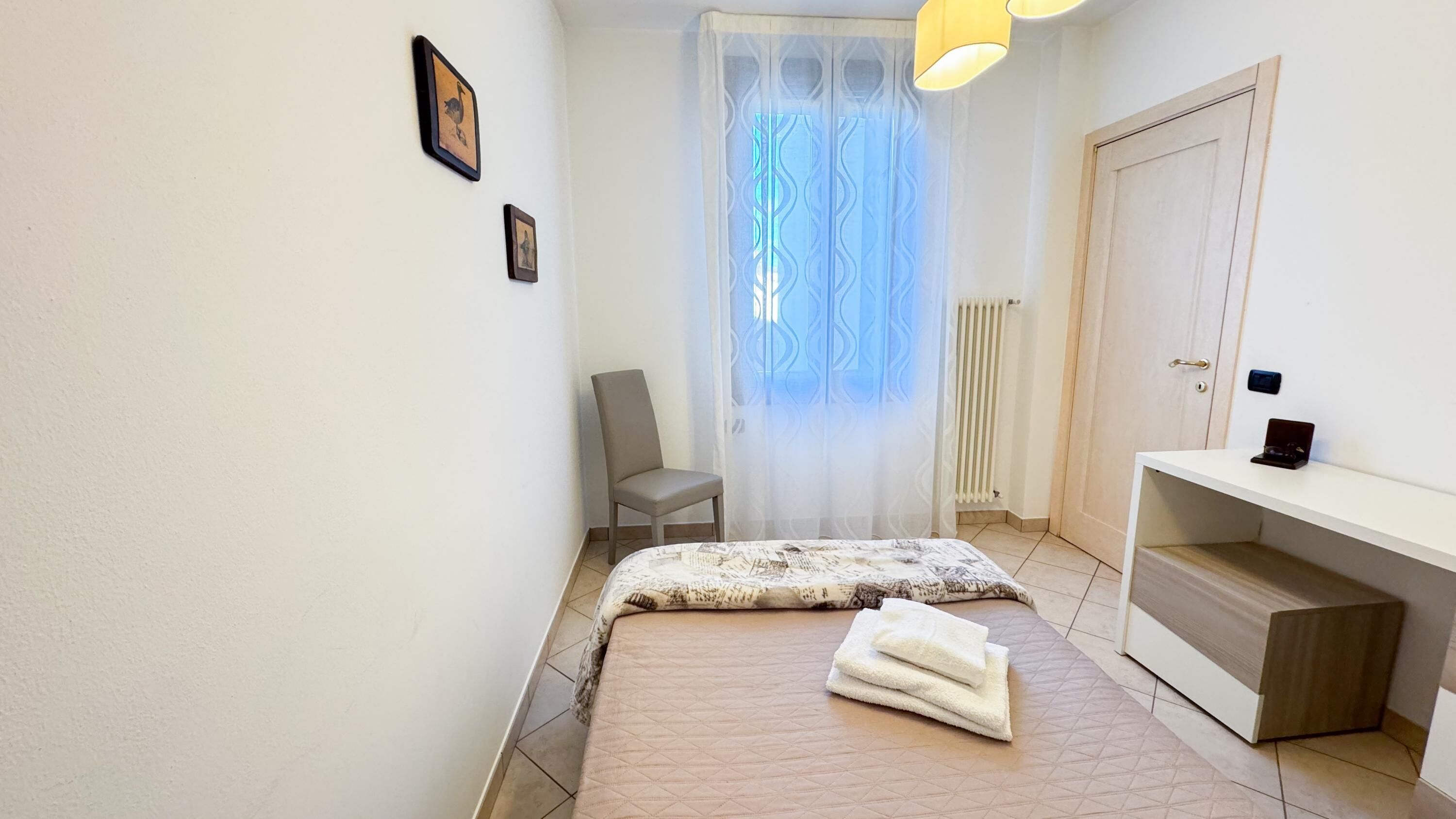 A bedroom with a bed, a chair, a bedside table, a radiator, and a window with sheer curtains.