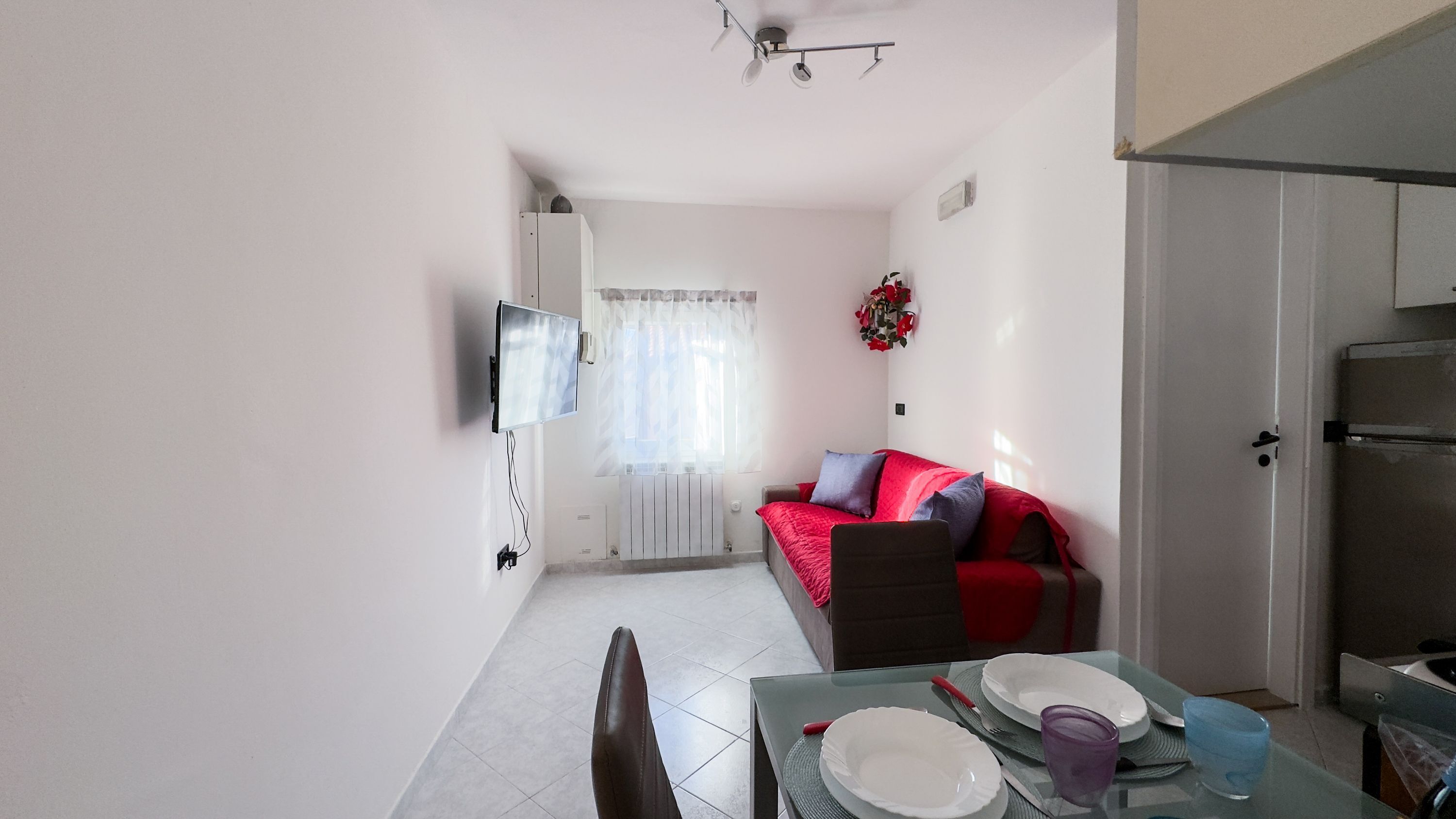 A compact living space with a dining area, a sofa with red and grey cushions, a TV mounted on the wall, and a kitchen area with a refrigerator.