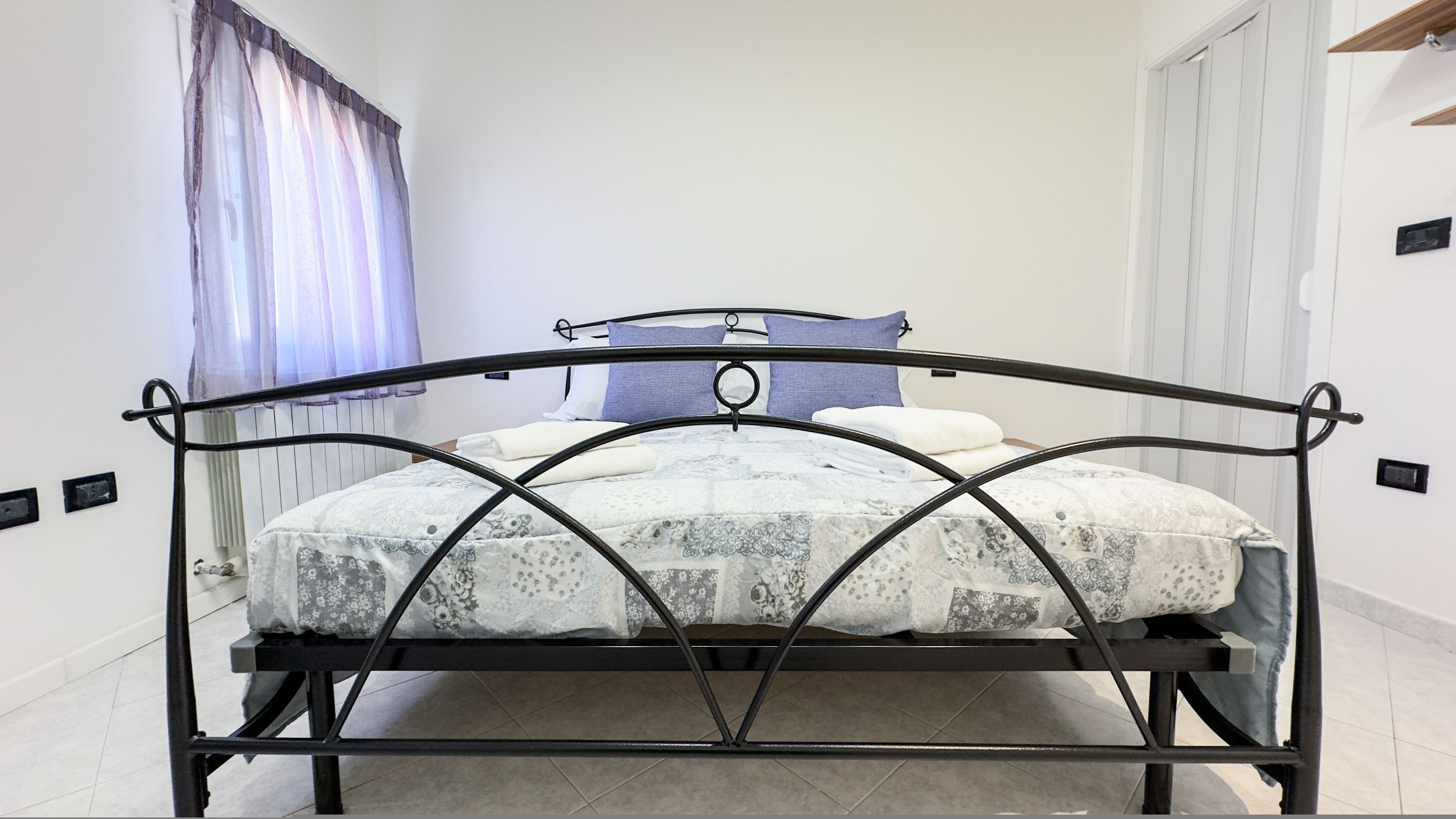Ein Bett mit Metallgestell, einer gemusterten Tagesdecke und einem blauen Kopfkissen.