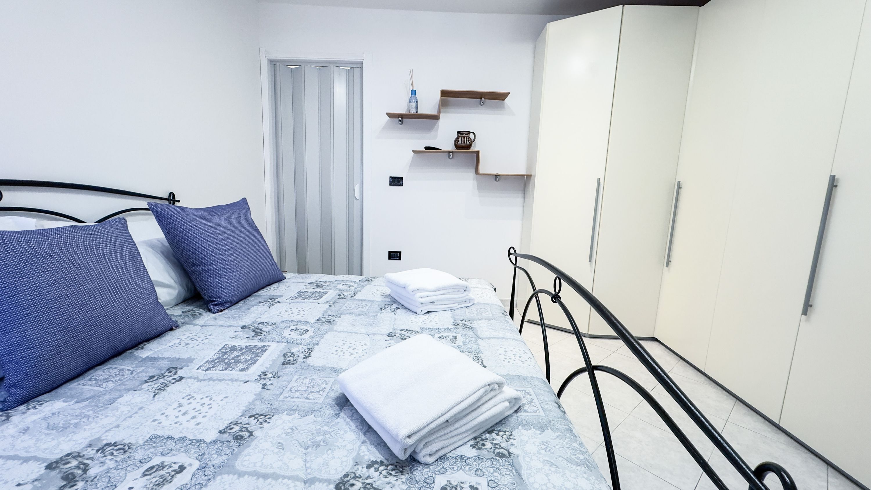 A bedroom with a bed, pillows, a wardrobe, and a small shelf with items.