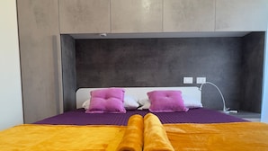 Quarto de hotel com cama, roupa de cama roxa, duas almofadas, cabeceira e tomada na parede.