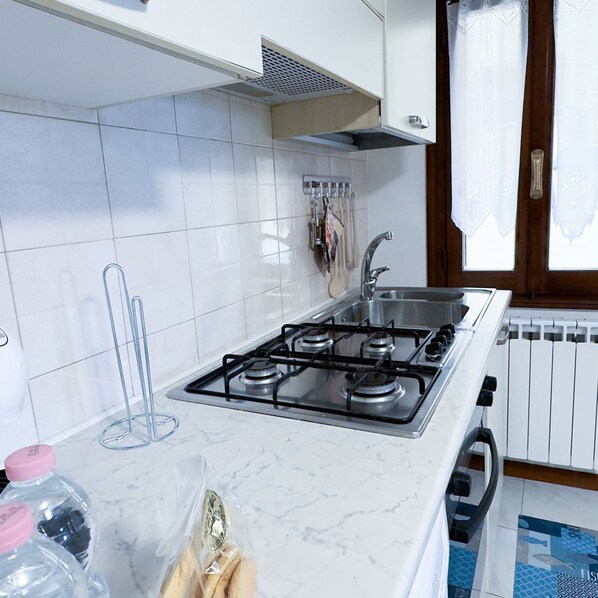 Una cocina con paredes de azulejos blancos, un lavamanos de acero inoxidable, una estufa a gas y una ventana con cortinas.