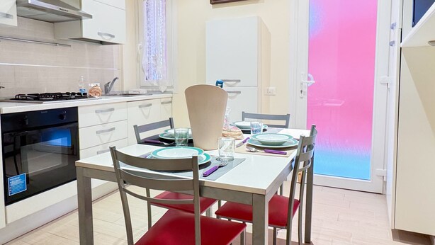 A modern kitchen with white cabinets, a gas stove, and a dishwasher. There is a dining table with red chairs and a painting on the wall. - Casa Dolce Casa by Ca-sa Check (Chioggia)