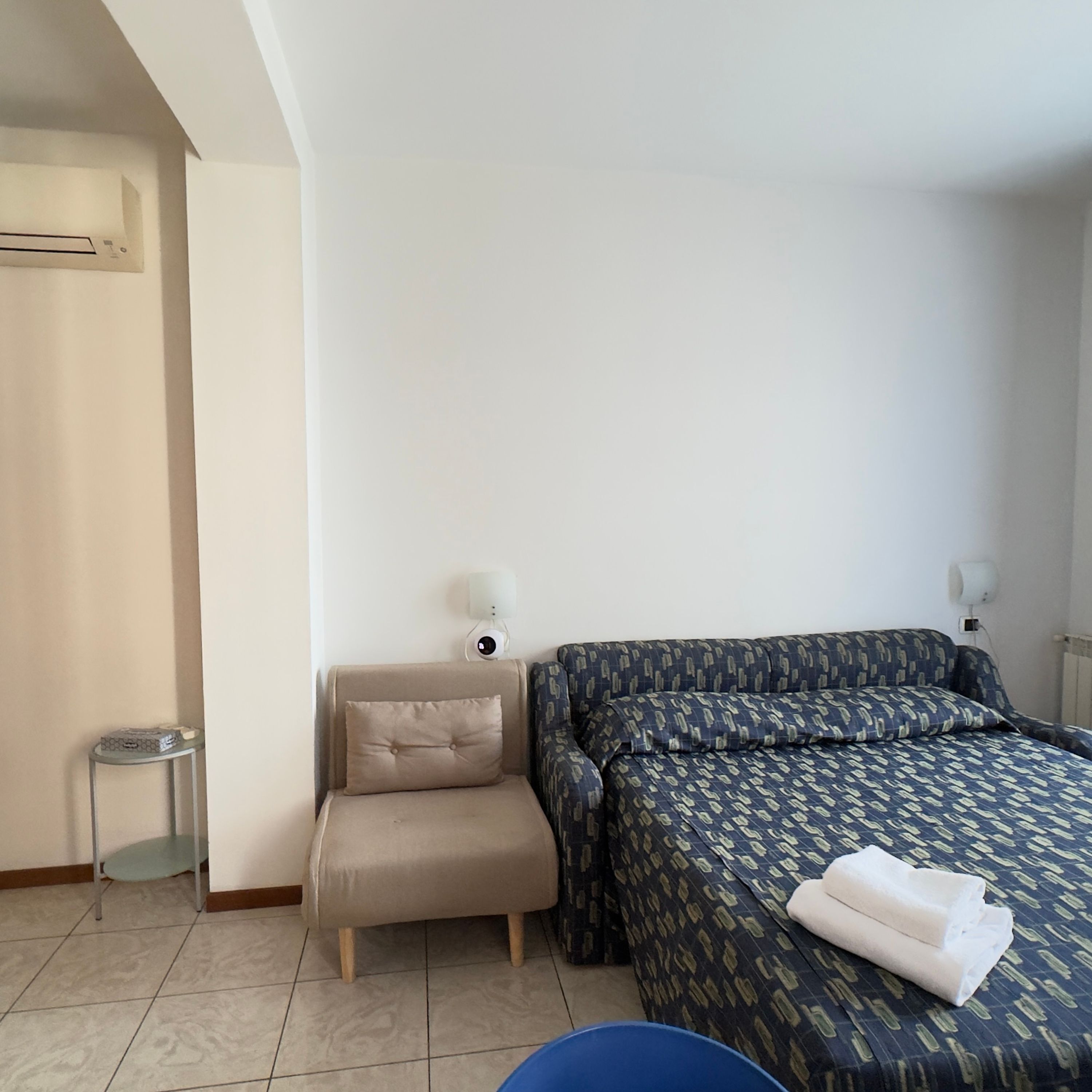 Hotellihuone, jossa on sänky, pieni nojatuoli, yöpöytä lampun kanssa ja patteri.