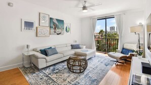 A modern living room with a white sofa, a round wicker coffee table, and a large window with a view of palm trees and buildings. - Palm Penthouse - Bright Designer Penthouse in the Heart of CityPlace (West Palm Beach)