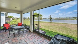 Um alpendre coberto com mesa e cadeiras, grandes janelas com vista para um lago e área gramada.