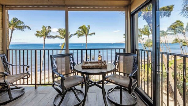 A balcony with a table and chairs, overlooking a beach with palm trees and the ocean. - Remodeled 2 bedroom oceanfront condo, AC, 6 mile sandy beach, shaved ice, bakery (Kihei)
