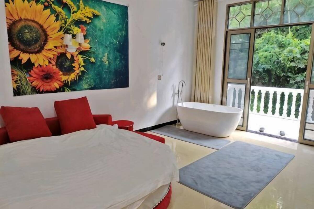 Ein Schlafzimmer mit einem großen Bett, einem Sonnenblumenbild, einer Badewanne und einem Balkon mit Blick auf Grünflächen.