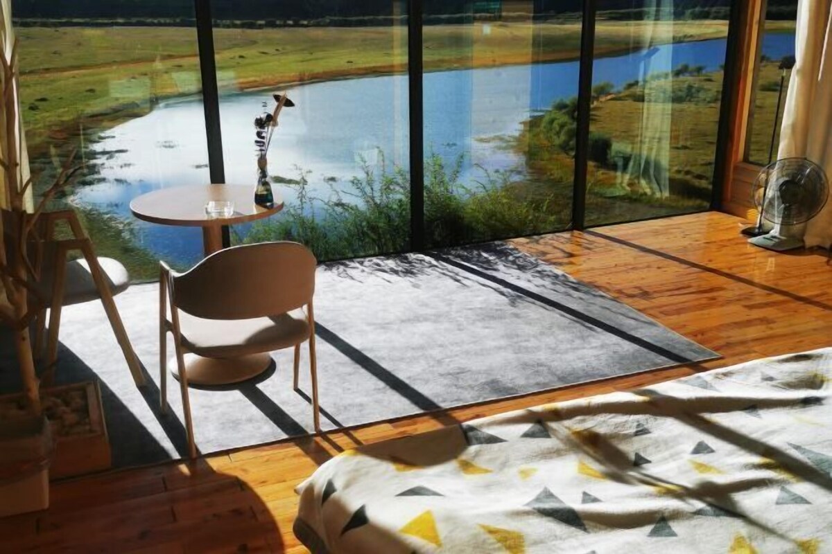 A bedroom with a large window overlooking a lake and fields, a bed, a chair, a small table, and a fan.