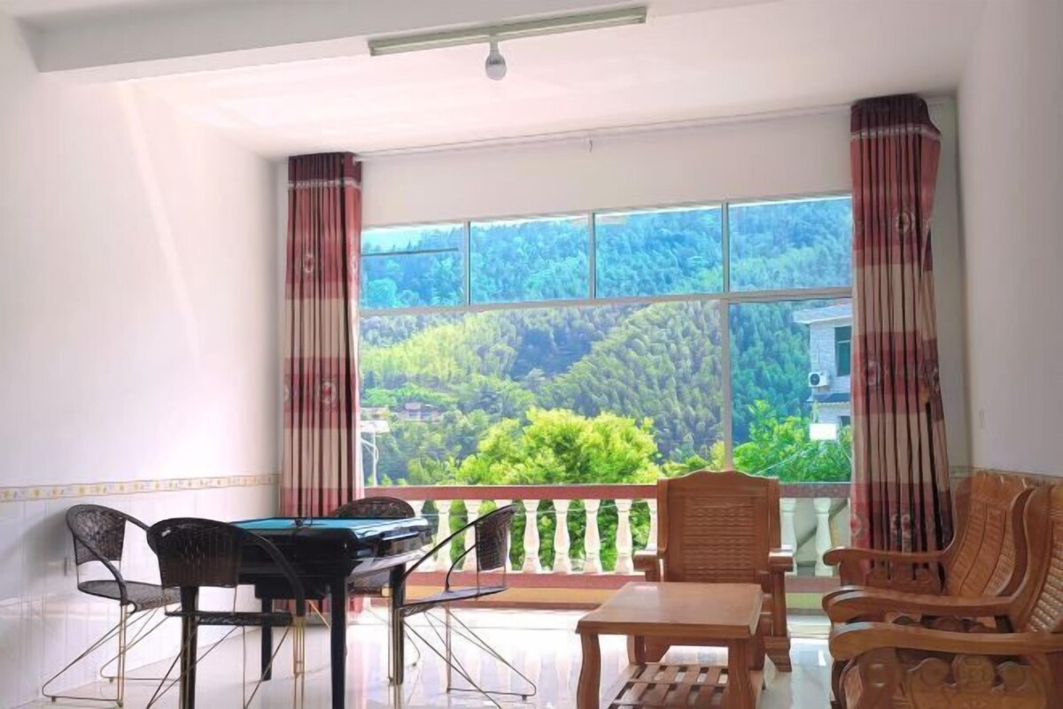 A room with a large window offering a view of a forested area. There is a pool table, a wooden table, and several chairs.