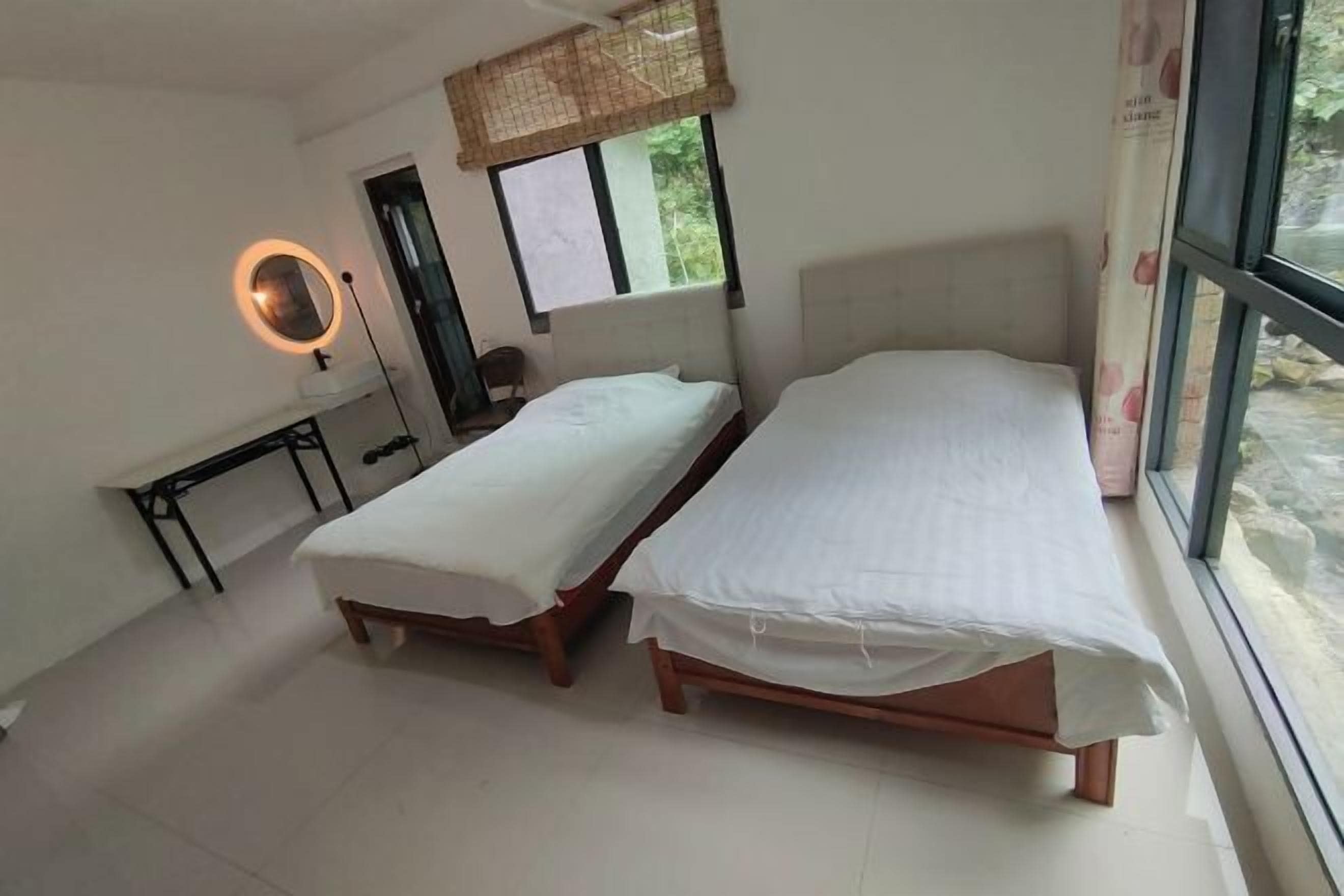 A room with two single beds, a bedside table, a mirror, and a window with a view of greenery.