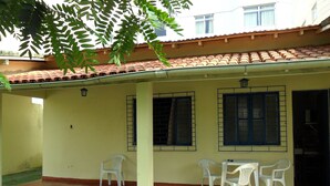 A covered patio area with a tiled roof, two windows with metal bars, and four white plastic chairs arranged around a small table.