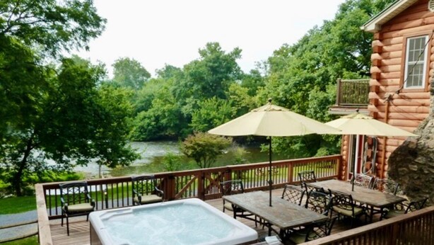 Une terrasse en bois avec un jacuzzi, du mobilier d’extérieur et une vue sur un lac entouré d’arbres.