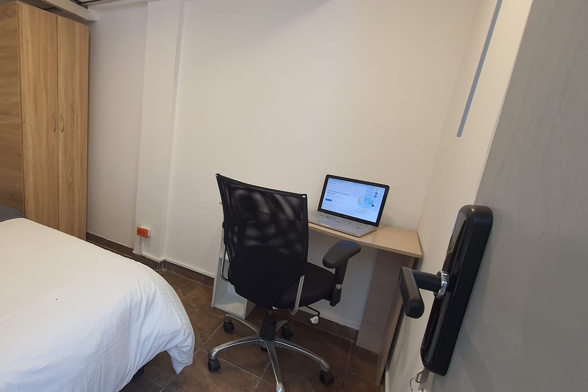 A bedroom with a bed, a wooden wardrobe, a desk with a laptop, and a chair.