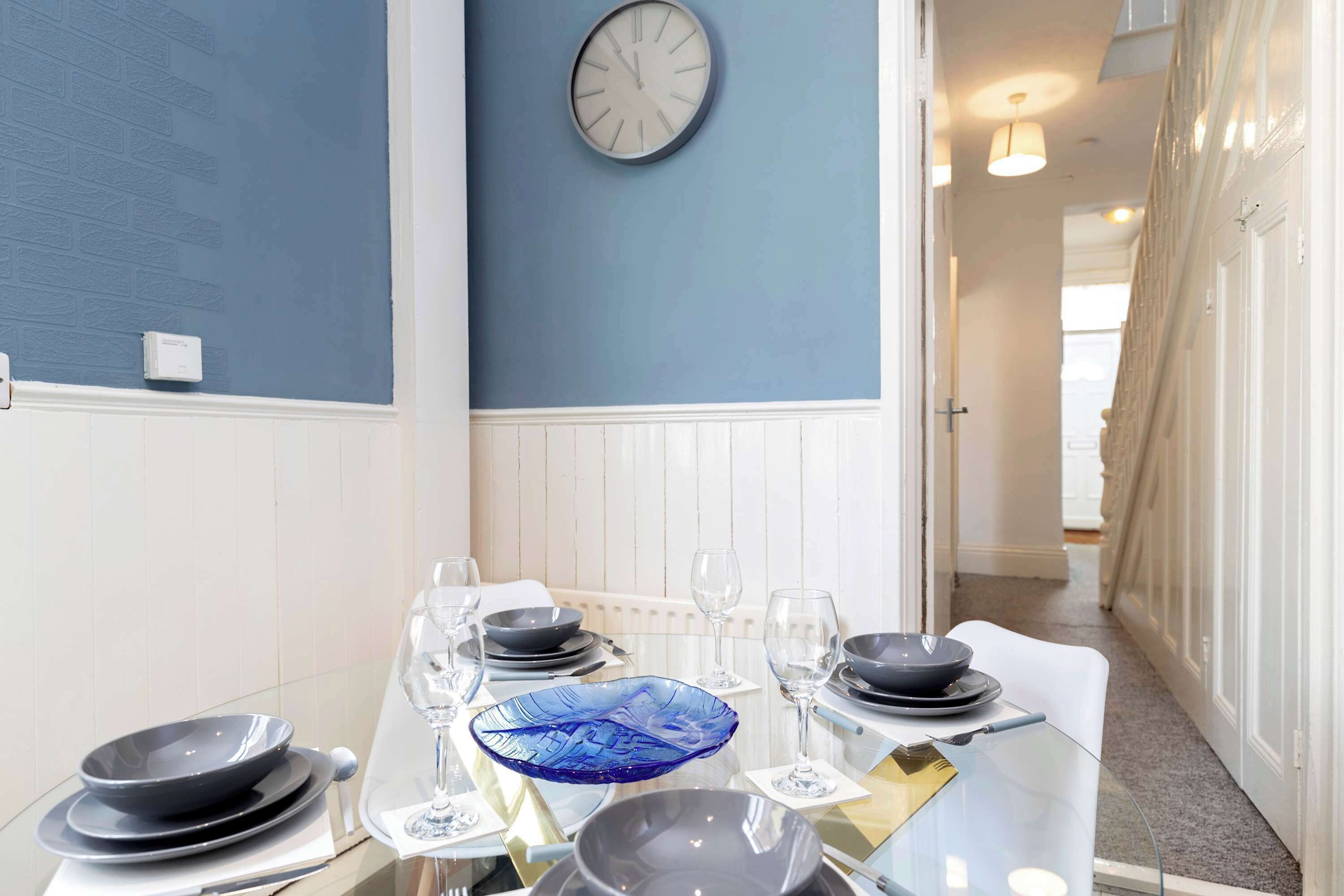 A dining area with a table set for four, a wall clock, and a hallway leading to another room.