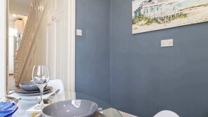 A dining area with a table set for two, a painting of a beach house on the wall, and a staircase leading to another room. - Large 4 Bedroom House Sleeps 9 With WiFi (Stockton-on-Tees)