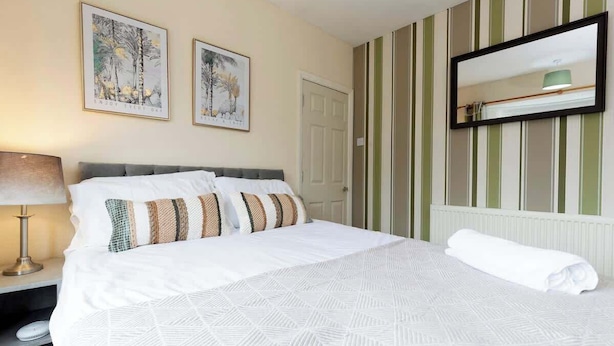 A hotel room with a bed, a nightstand, a lamp, two framed pictures on the wall, a door, and a mirror. - Comfortable 3 Bedroom Home Sleeps 5 (Stockton-on-Tees)