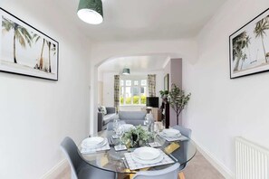 A dining area with a glass table set for four, surrounded by grey chairs. There are framed pictures on the walls, a plant on the table, and a view into another room through an archway. - Comfortable 3 Bedroom Home Sleeps 5 (Stockton-on-Tees)