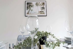 A dining table set with clear glassware, a potted plant, and a framed picture on the wall.