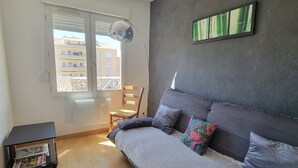 Une chambre avec un canapé-lit, un fauteuil, une petite table avec des journaux, une fenêtre avec des stores et un tableau au mur.