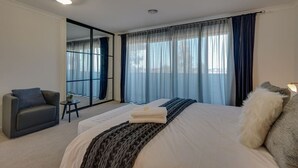 A hotel room with a bed, a chair, a small table, and a window with curtains.