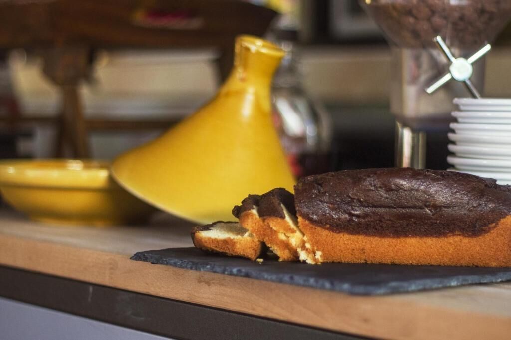 Un pastel de chocolate sobre una tabla de pizarra con un jarrón amarillo y una máquina de café al fondo.