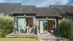 A wooden cottage with two open front doors, a small patio with a table and chairs, and a garden area.
