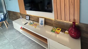 Een moderne woonkamer met een grote flatscreen televisie aan een houten wand, een witte tv-kast eronder en een kleine rode vaas aan de rechterkant.