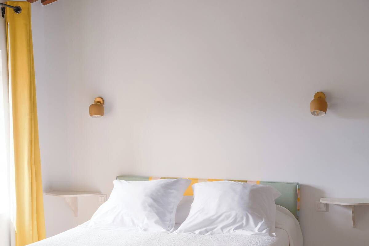 Un lit avec du linge de lit blanc, deux appliques murales et un rideau jaune.