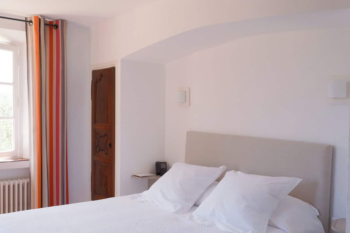 Une chambre à coucher avec un lit, du linge de lit blanc, une porte en bois et une fenêtre avec des rideaux.