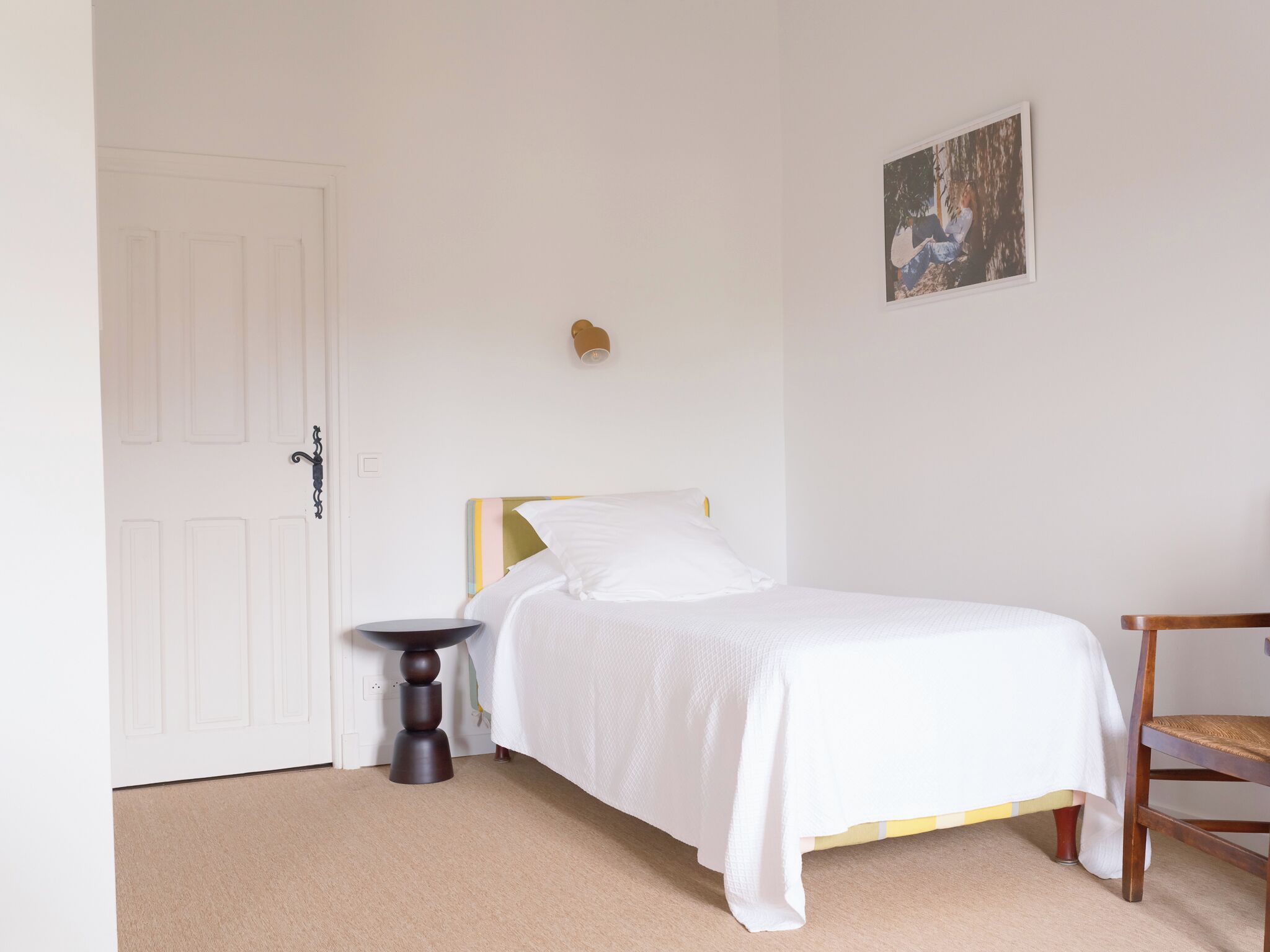 A bedroom with a bed, a small round table, a chair, a door, and a wall-mounted light fixture.