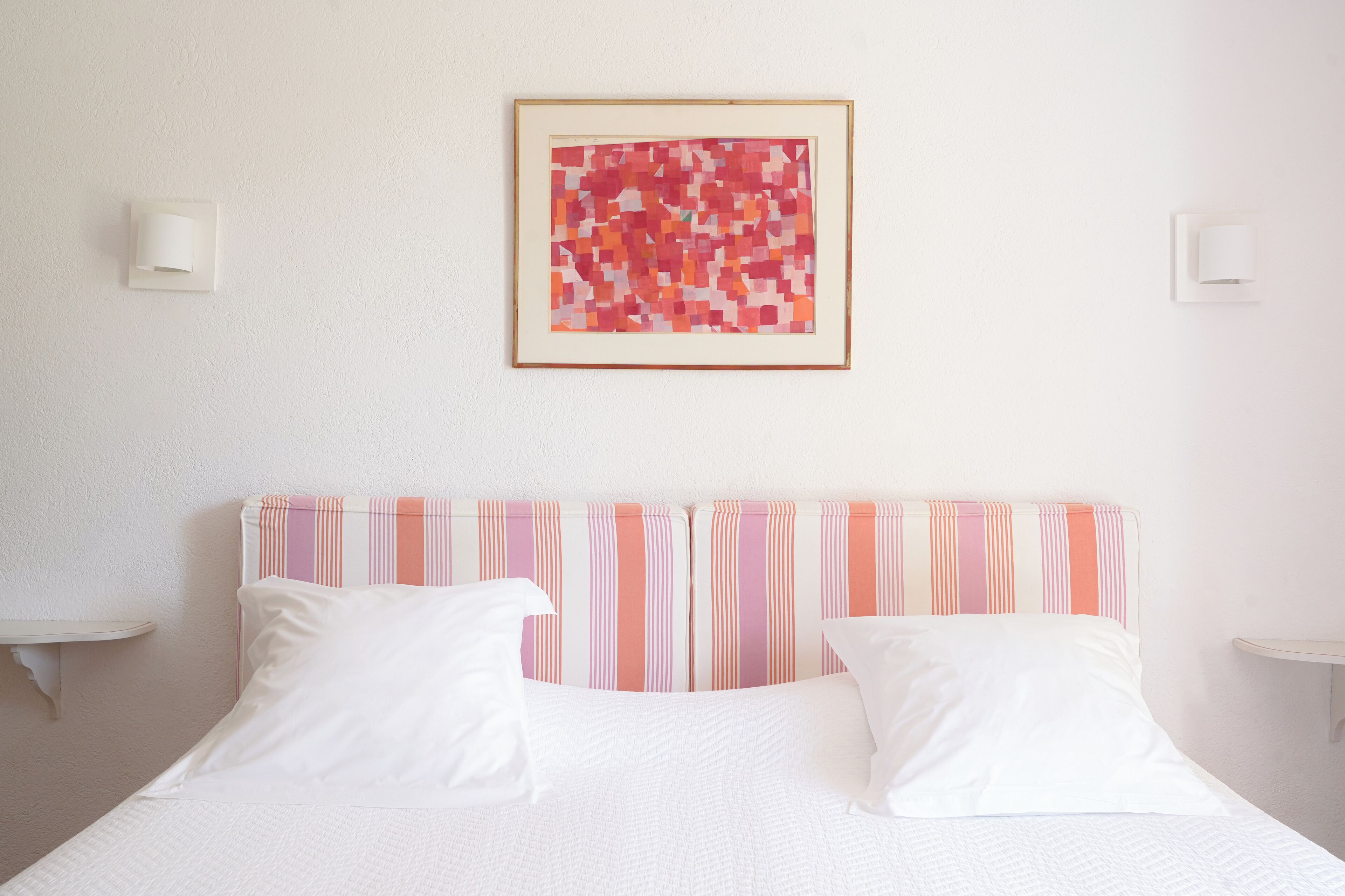 Een net opgemaakt bed met witte lakens, een gestreept hoofdbord en een ingelijst abstract schilderij aan de muur.