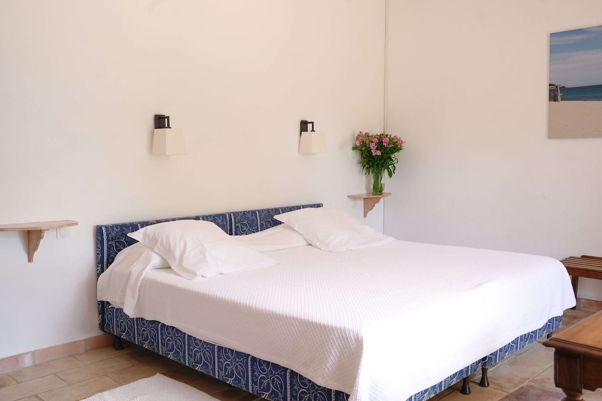 Un lit bien fait, avec une tête de lit bleue, deux lampes fixées au mur, une petite étagère en bois et un tableau encadré accroché au mur.