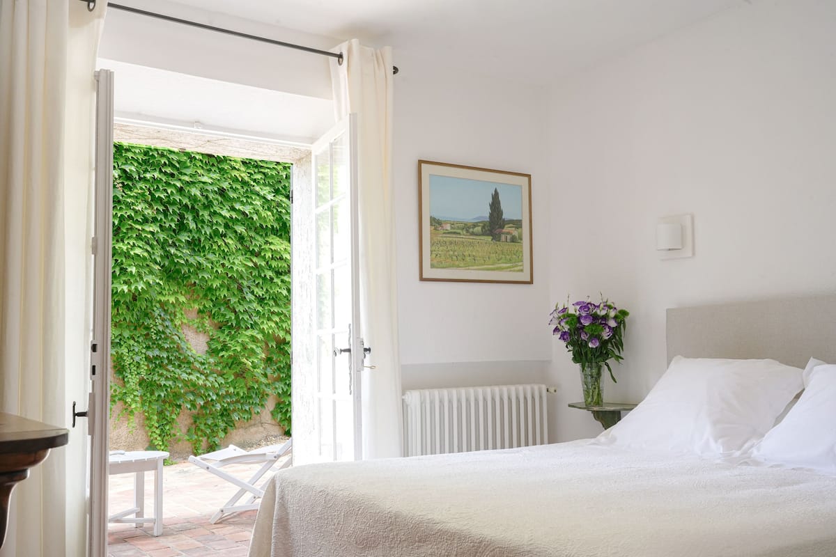 Une chambre à coucher avec un lit, un radiateur blanc, un tableau accroché au mur et une vue sur un jardin, visible à travers une porte ouverte.